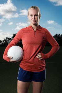 This is a portrait of a 15 year old teenage girl holding a soccer ball out on a field. She is looking seriously into the camera with a tough competitive spirit.